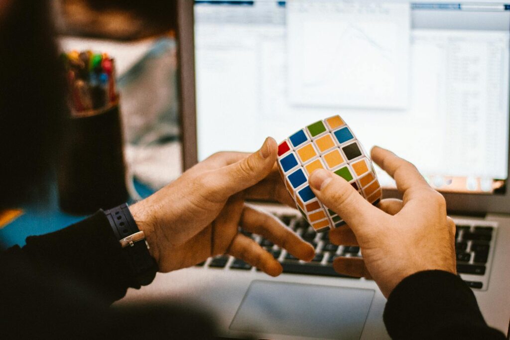 Close-up of hands solving a Rubik's Cube in front of a laptop showcasing a relaxed work environment. mãos tentando resolver um cubo mágico